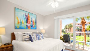 Habitación de hotel con una cama grande, un cuadro de veleros, un balcón con vistas a palmeras y edificios, y un ventilador de techo.