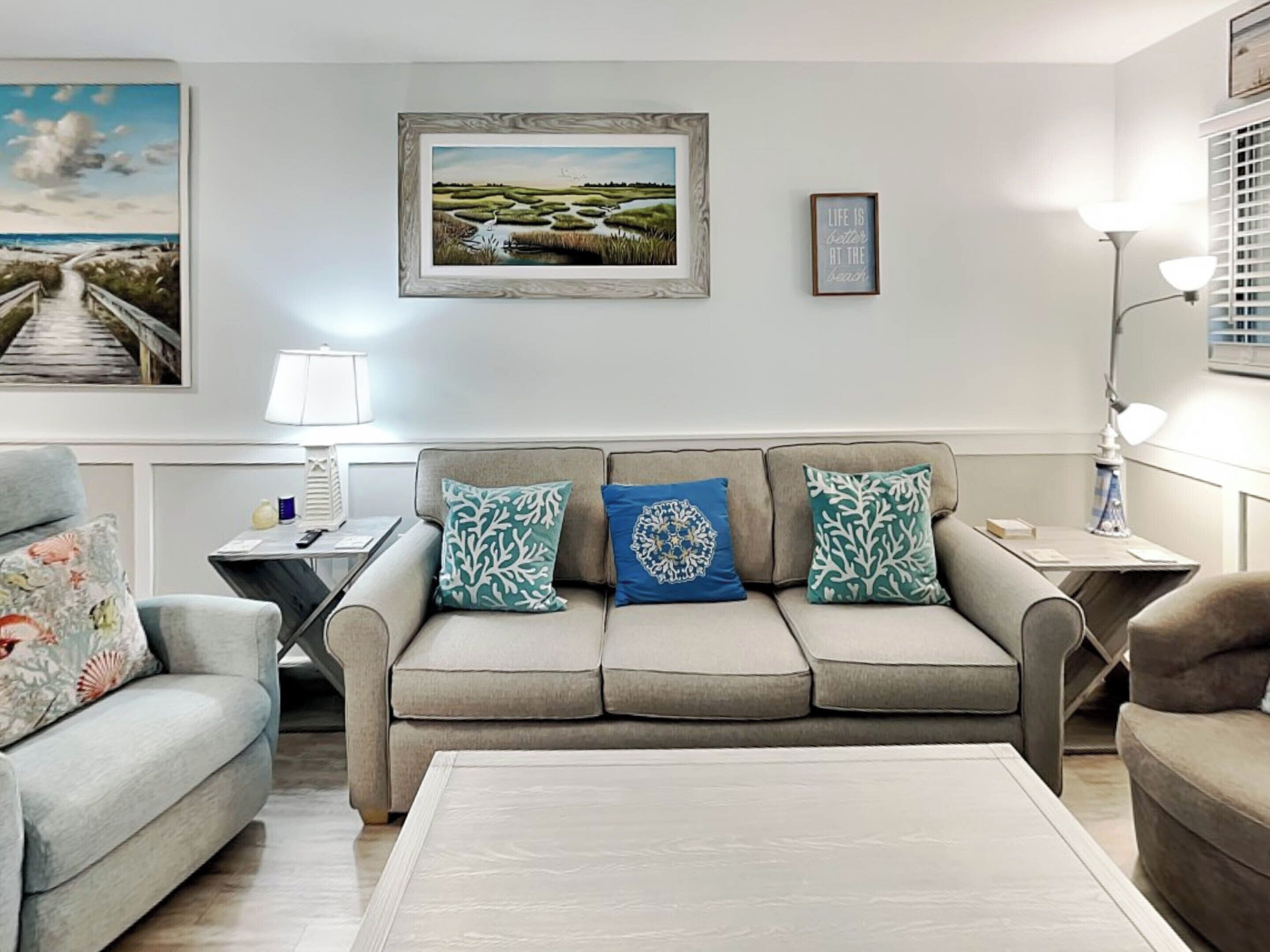 A living room with a beige sofa, two patterned throw pillows, a coffee table, and a painting of a beach scene.