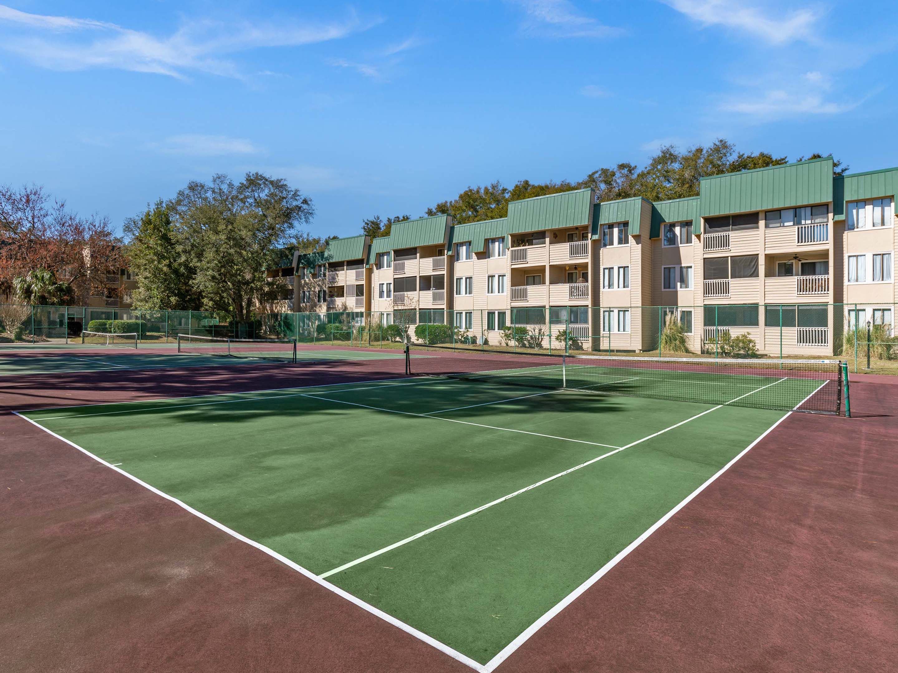 A tennis court with a net, surrounded by a fence, and adjacent to a multi-story building with balconies.