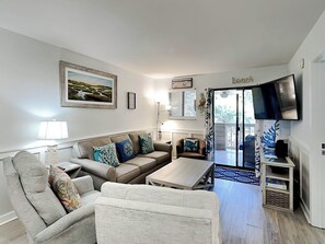 A living room with a sofa, armchairs, a coffee table, a TV, and a painting on the wall. - The Spa 1219 by Hilton Head Retreats (Hilton Head Island)