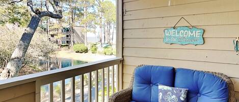 A porch with a wicker sofa, a 'Welcome to the Beach' sign, and a view of a swimming pool and buildings.