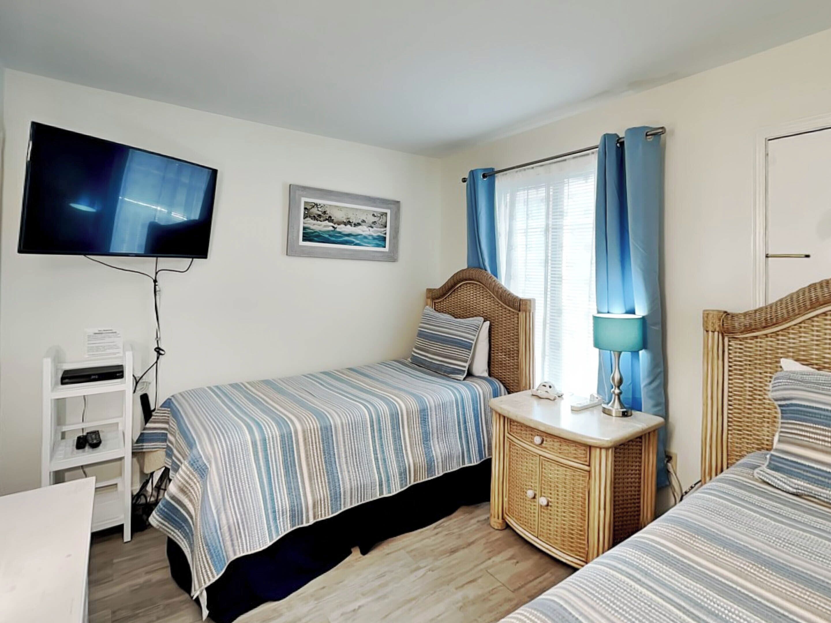 Ein Schlafzimmer mit zwei Betten, einem Fernseher, einem Bild an der Wand und einem Fenster mit Vorhängen.