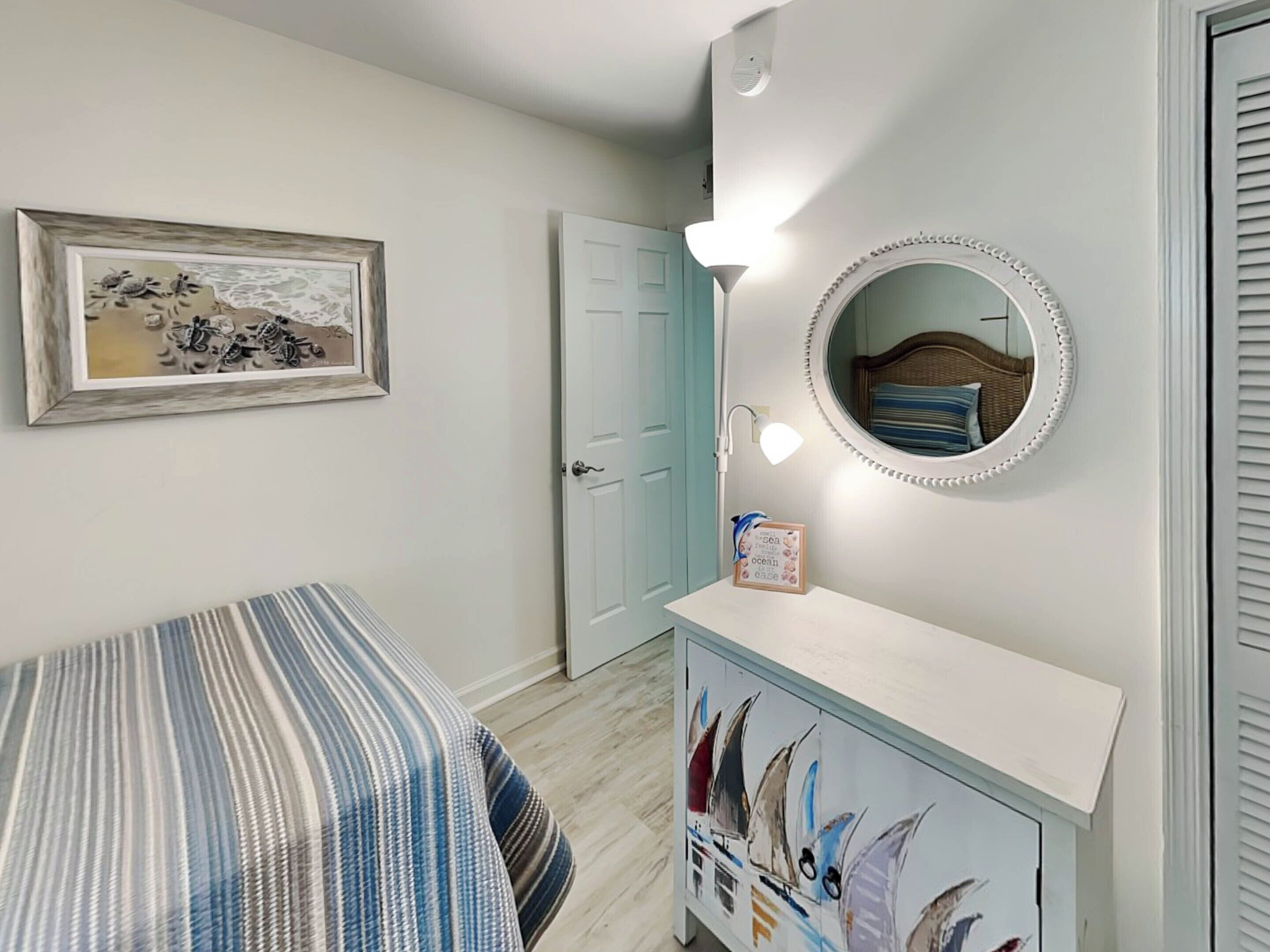 Une chambre à coucher avec un lit, une commode avec un miroir, un tableau encadré au mur et une porte.