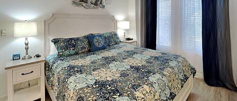 Une chambre à coucher avec un grand lit, deux tables de chevet, une commode, un élément décoratif mural et une fenêtre avec des rideaux.