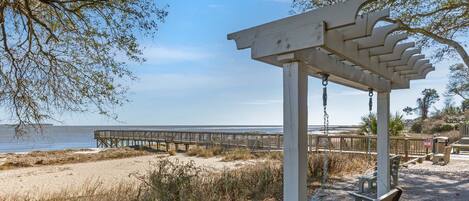 Une pergola en bois avec une balançoire, donnant sur une plage où l’on aperçoit, au loin, une jetée en bois.