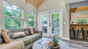 A cozy living room with a green sofa, a coffee table with decorative items, large windows with a view of trees, and a sliding glass door leading to an outdoor area.