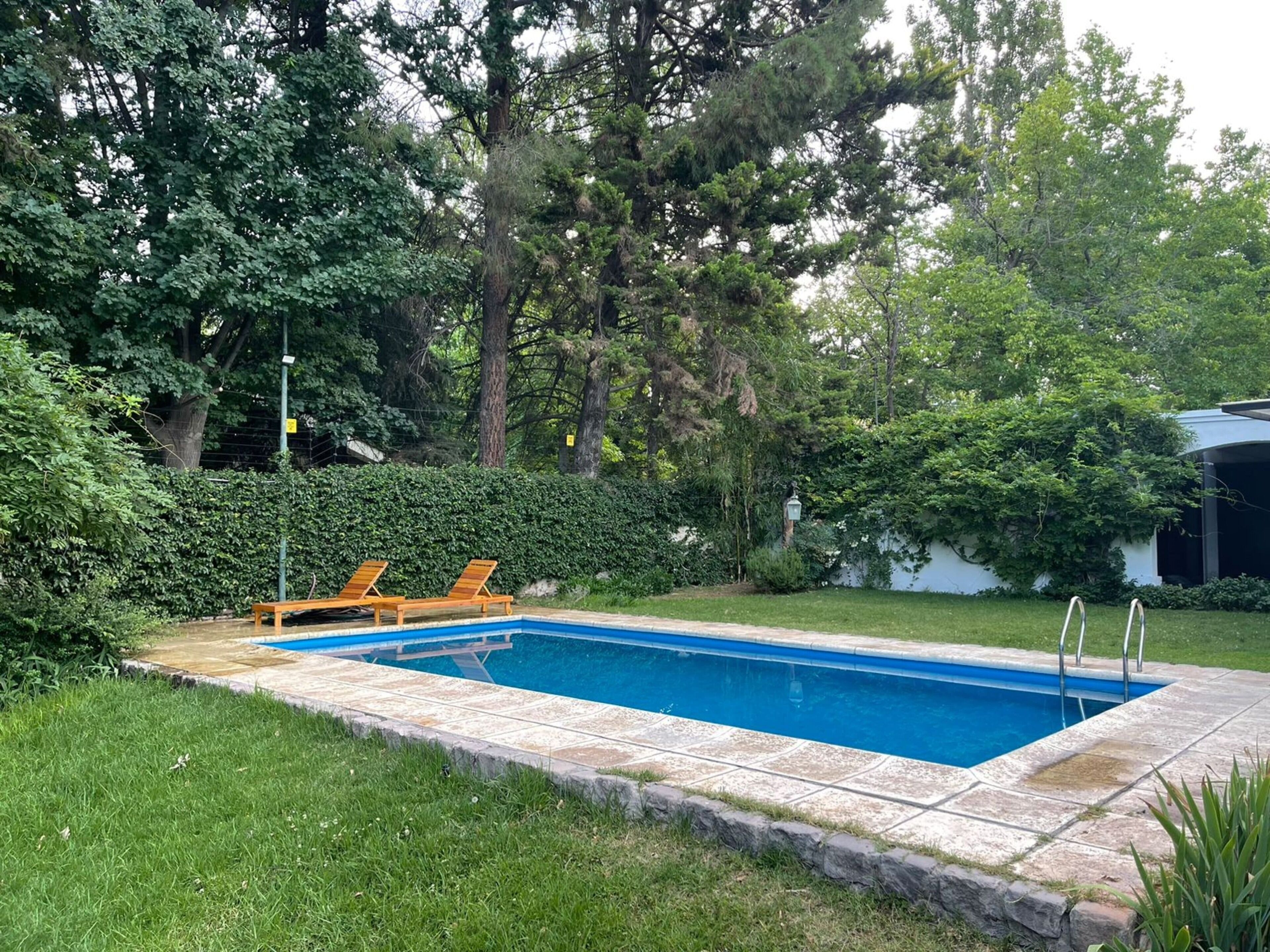 A rectangular swimming pool with a tiled deck, surrounded by lush greenery and trees.