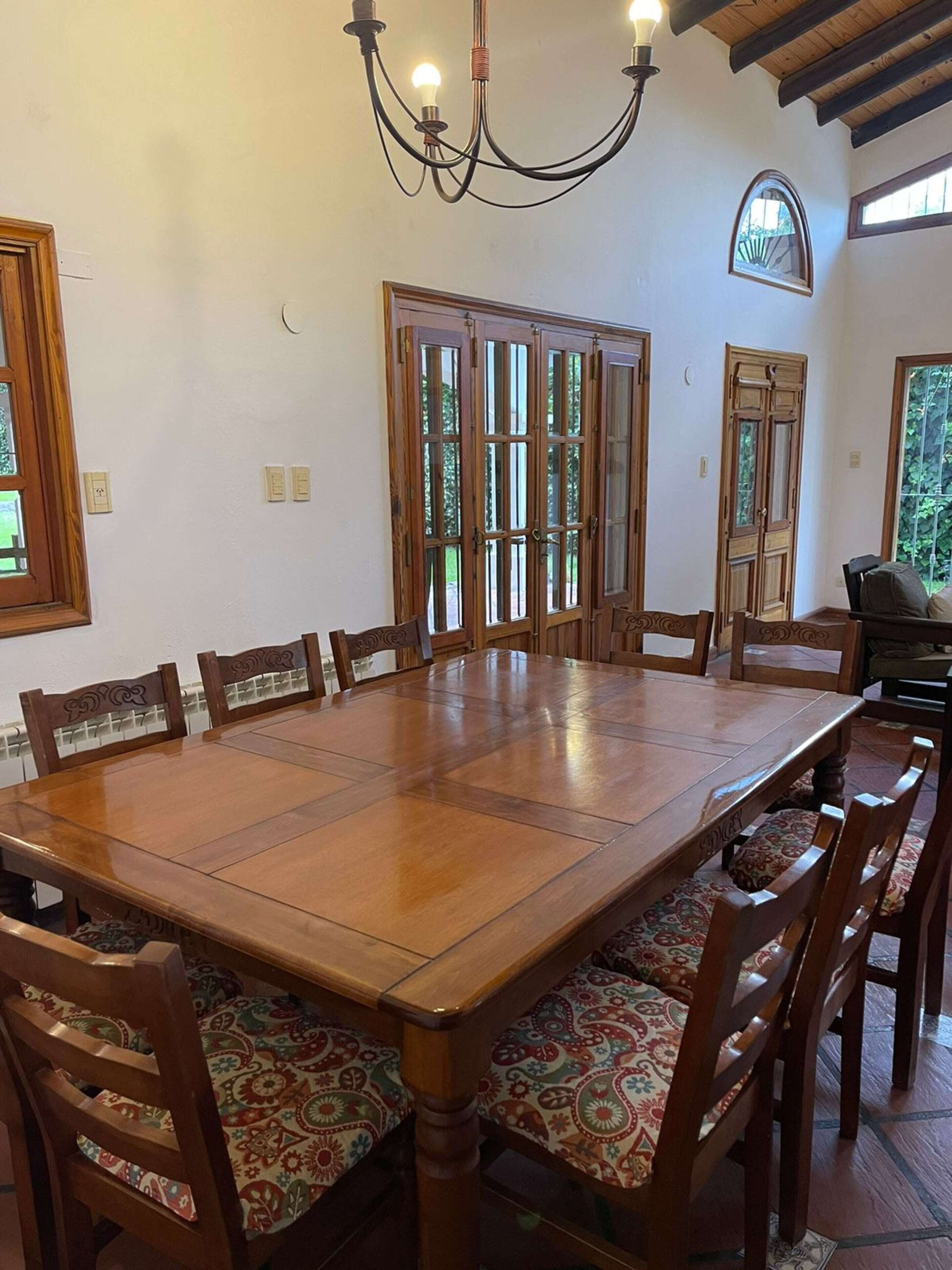 Een eetkamer met een grote houten tafel, omringd door stoelen met gestreepte kussens, een kroonluchter en uitzicht op groen buiten.