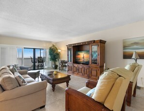 A spacious living room with a large TV, comfortable seating, a dining area, and a balcony with a view. - Reflections on the Gulf by Distinctive Beach Rentals (Indian Rocks Beach)