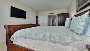 A bedroom with a large bed, a wooden headboard, a flat-screen TV mounted on the wall, and a door leading to another room. - Reflections on the Gulf by Distinctive Beach Rentals (Indian Rocks Beach)