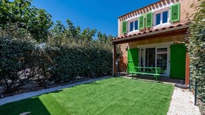 Een huis met een verzorgd gazon, een terras met een bank en een deur met groene luiken.