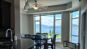 A kitchen with a large island, bar stools, a dining table with chairs, a ceiling fan, and large windows with a view of a lake and mountains. - Sunset Waterfont Resort 12. Luxury Lakeside, Lakeview 3 bed/2bath Kelowna Condo. (Kelowna)