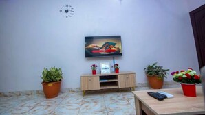 A living room with a flat-screen TV mounted on the wall, a wooden cabinet with decorative items, and potted plants. - AJI (1.0) | Warm 2BED Apartment (Ijegun, Lagos) (Lagos)