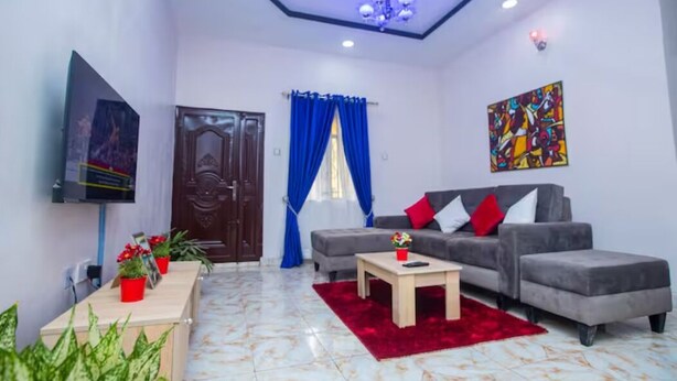 A modern living room with a grey sofa, red and white cushions, a wooden coffee table, and a red rug. There is a TV mounted on the wall, a door with a wooden frame, and a blue curtain. - AJI (1.0) | Warm 2BED Apartment (Ijegun, Lagos) (Lagos)