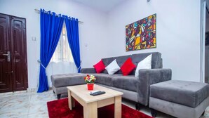A living room with a grey sofa, red and white cushions, a wooden coffee table with a vase of flowers, and a red rug. - AJI (1.0) | Warm 2BED Apartment (Ijegun, Lagos) (Lagos)