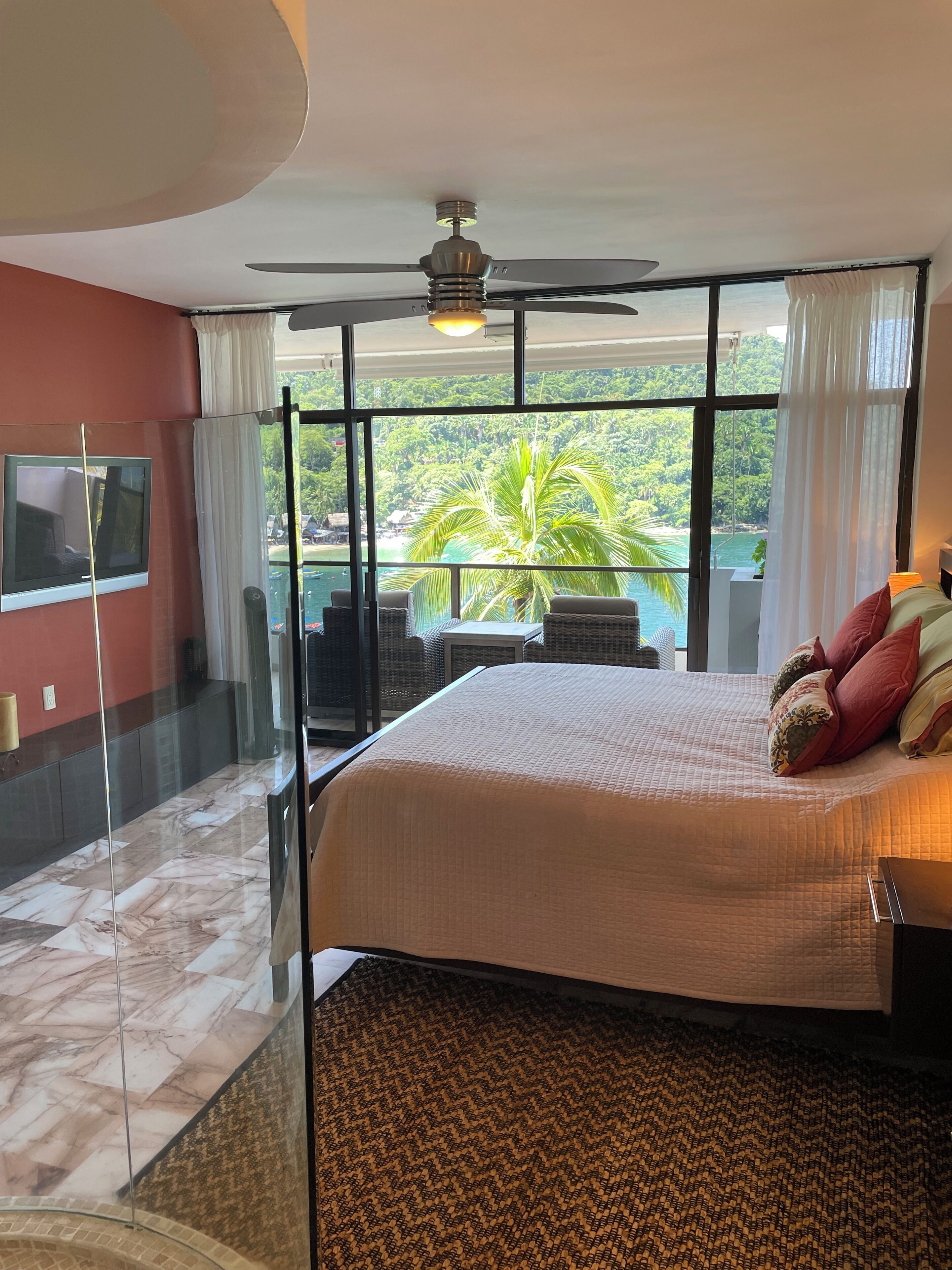 Ein Hotelzimmer mit Bett, Deckenventilator, großem Fenster mit Meerblick und Fernseher an der Wand.
