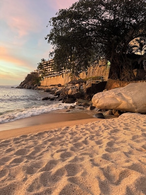 A beach with a rocky shoreline, a building with balconies overlooking the sea, and a thatched-roof structure.
