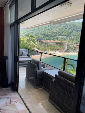 A balcony with wicker furniture, a view of a beach, and a building in the background.