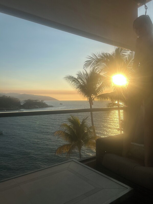 Vista desde un balcón sobre un cuerpo de agua, con una palmera y montañas a lo lejos.