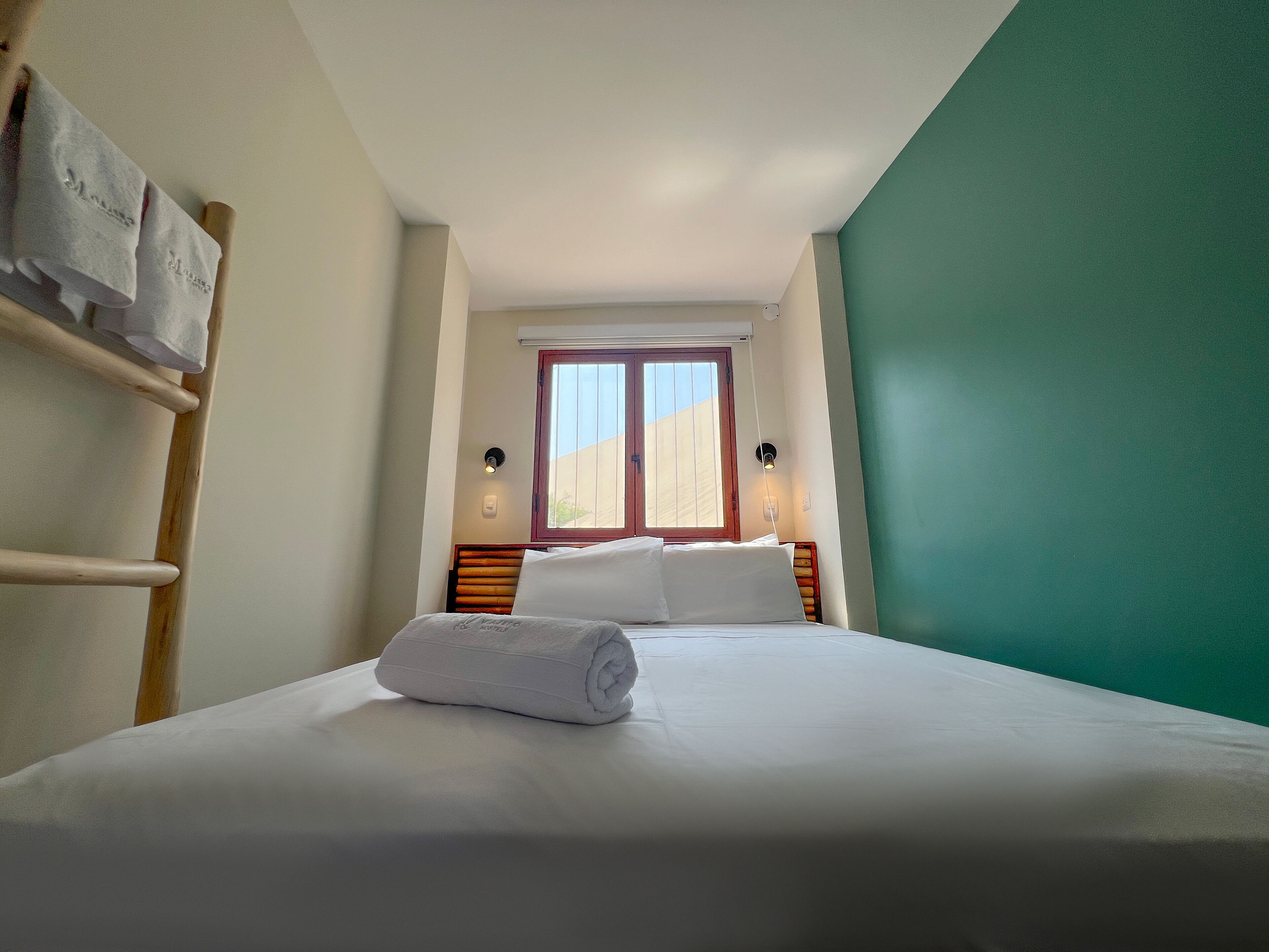 Una habitación de hotel con una cama, una ventana, un perchero en la pared y una lámpara de pared.