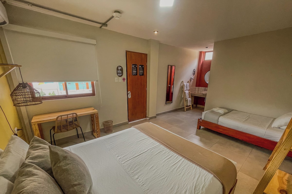A hotel room with a large bed, a desk, a chair, a small table, a window, and a bathroom with a sink and mirror.