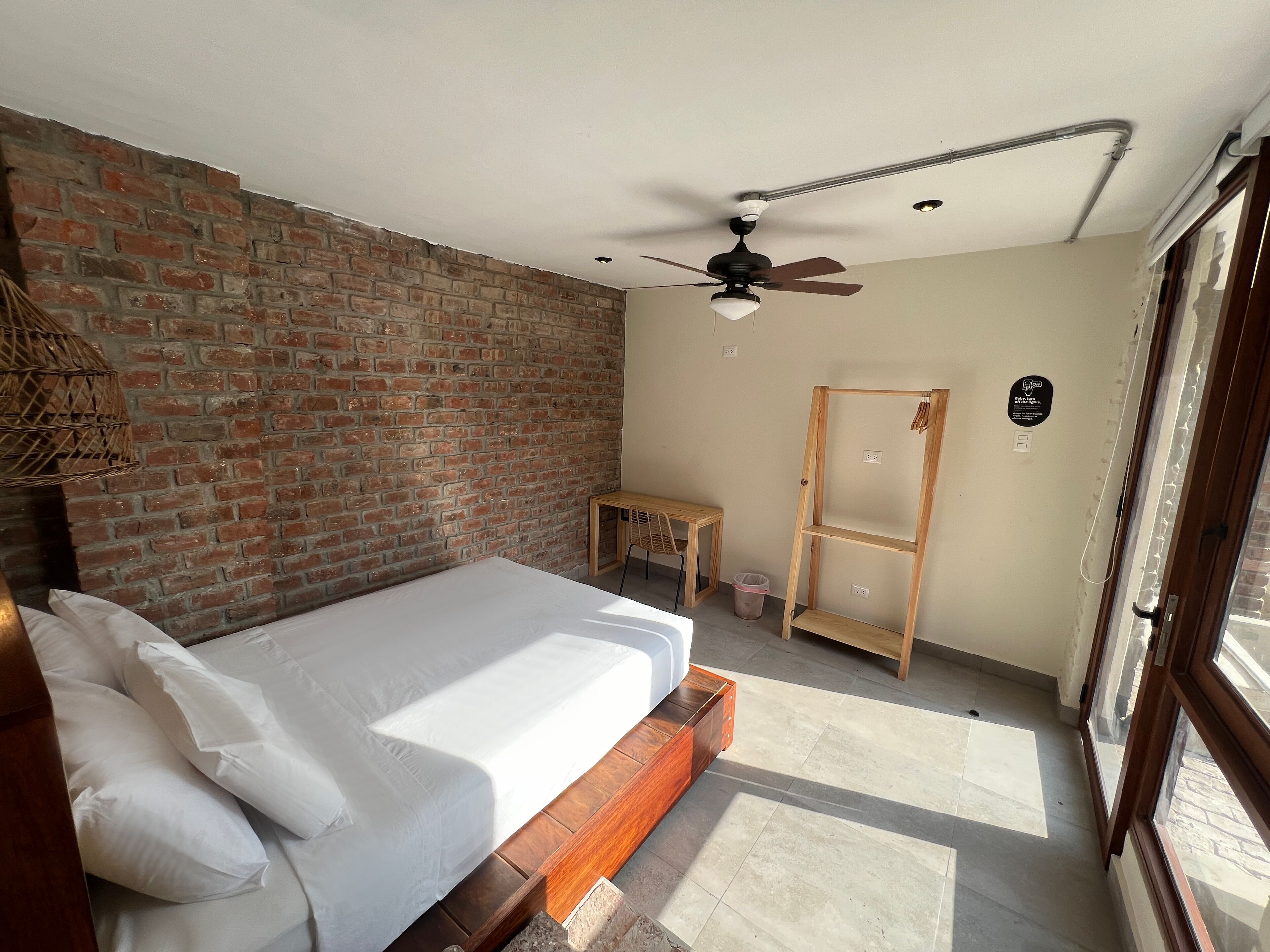 A bedroom with a brick wall, a bed with white linens, a ceiling fan, a wooden nightstand, a small table, and a window with blinds.