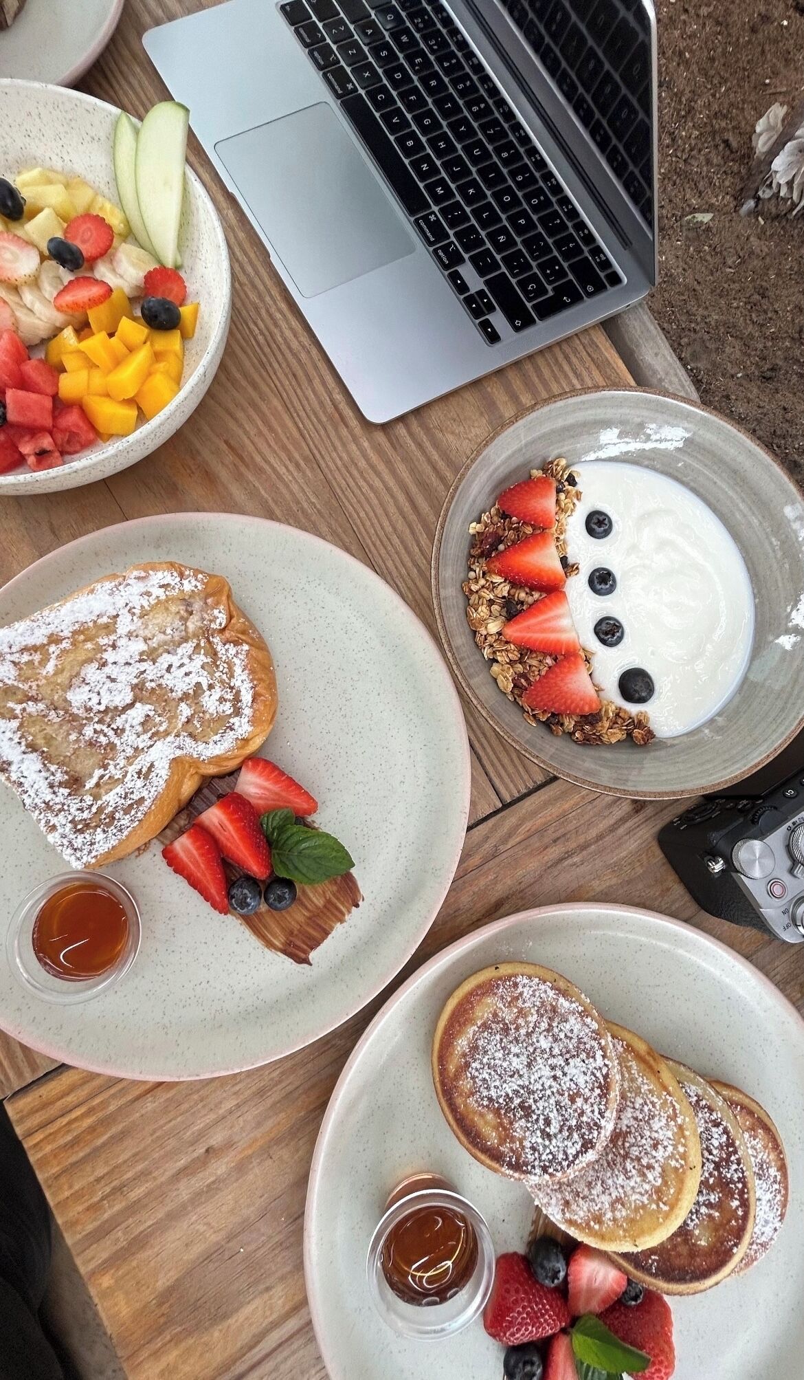 Een ontbijtje met pannenkoeken, fruit, yoghurt en een laptop op een houten tafel.