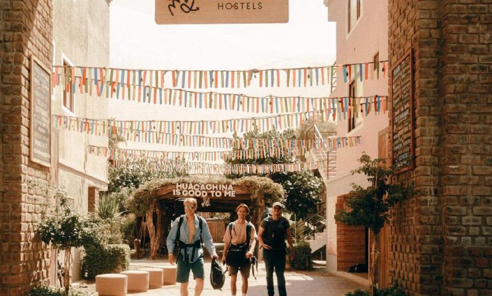 Une cour d’auberge avec une pancarte sur laquelle on peut lire : « HUACAGHINA ME PLAÎT », entourée d’immeubles en briques et de drapeaux aux couleurs vives.