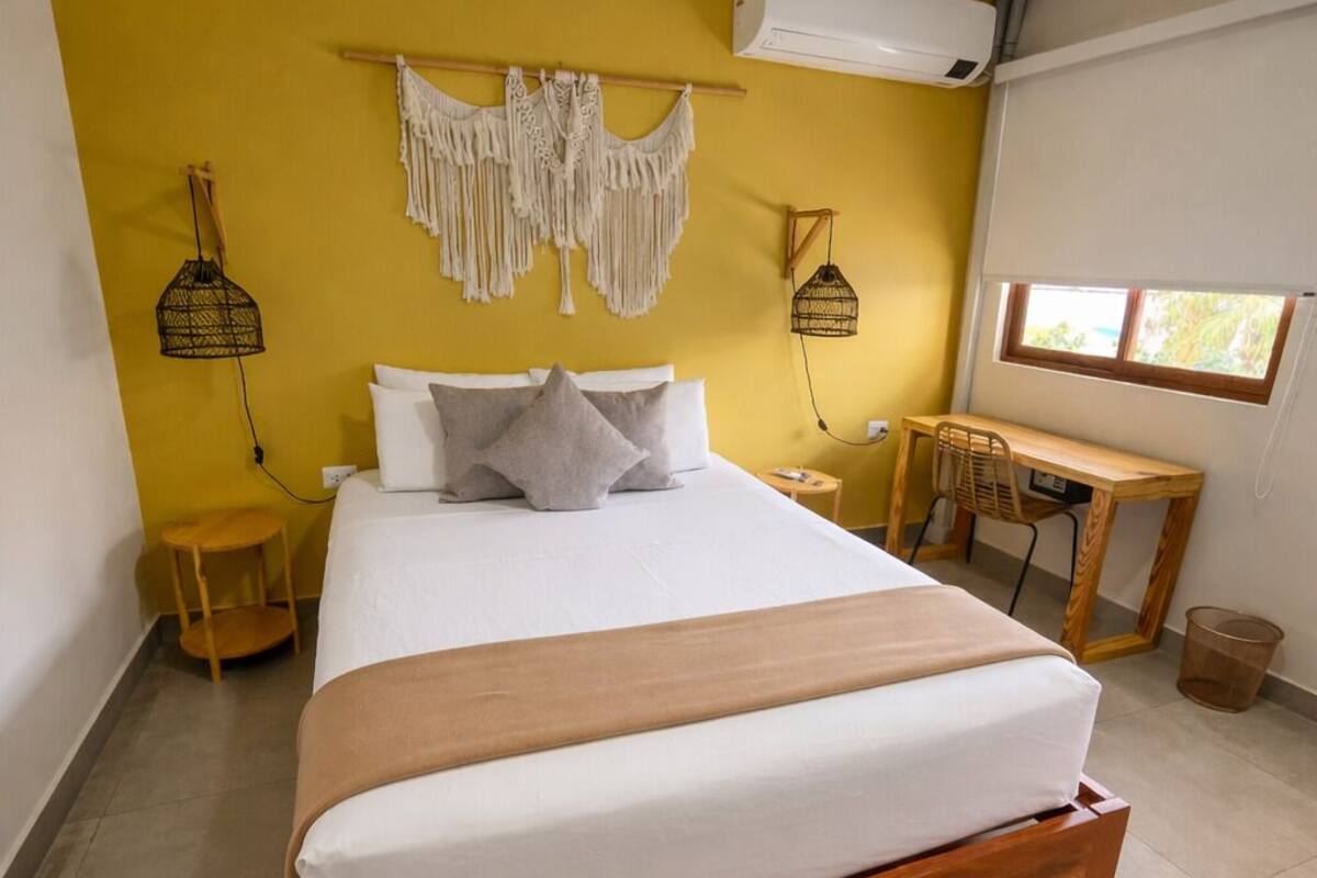 A bed with a wooden frame, a desk, a chair, a macrame wall hanging, and an air conditioning unit.