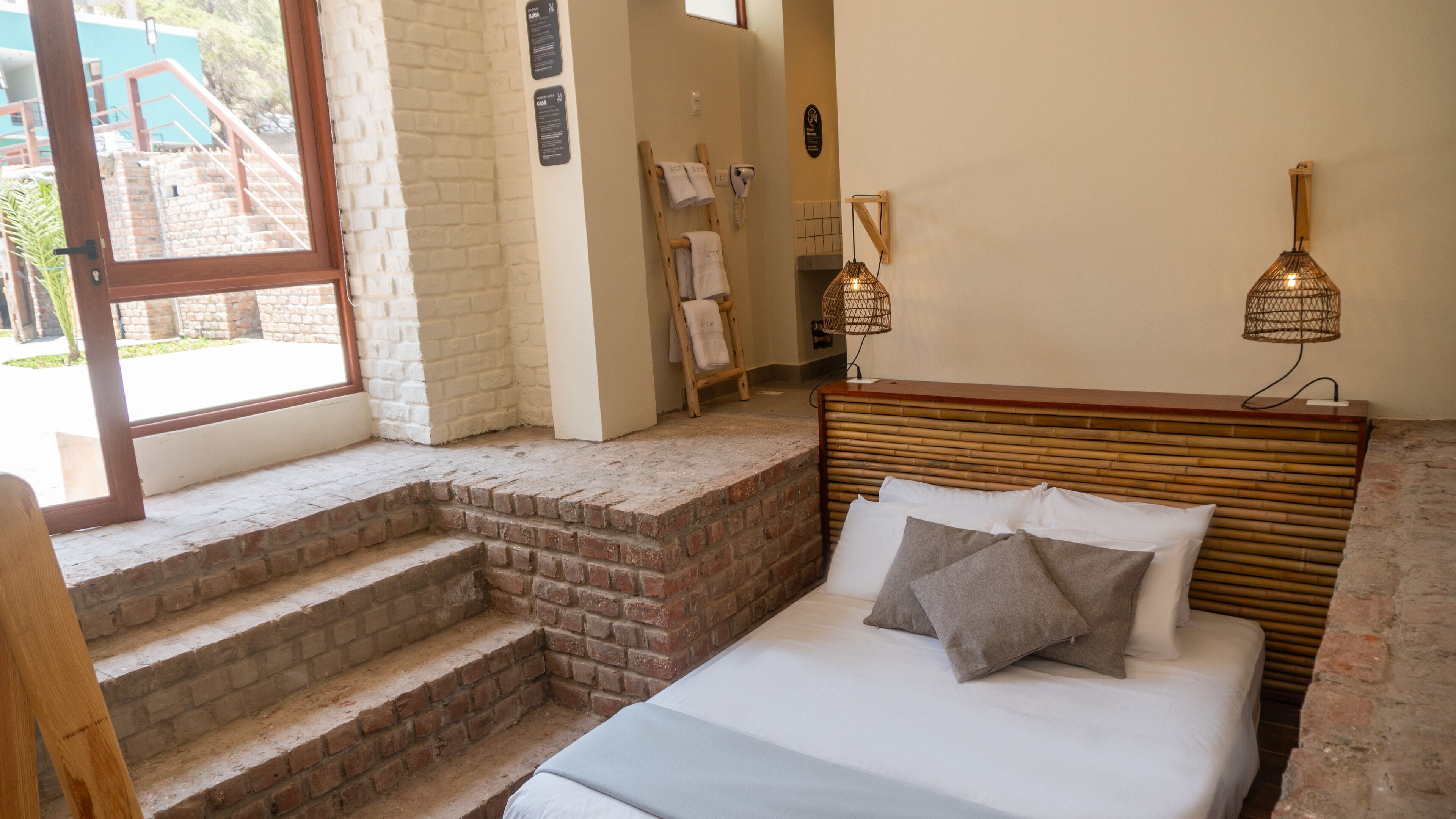 Un dormitorio con cama, cabecera de madera, pared de ladrillo, ventana y un pequeño tramo de escaleras que conducen a una puerta.
