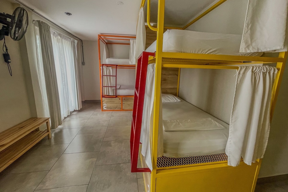 A dormitory room with bunk beds, a wooden bench, a fan, and a curtain.