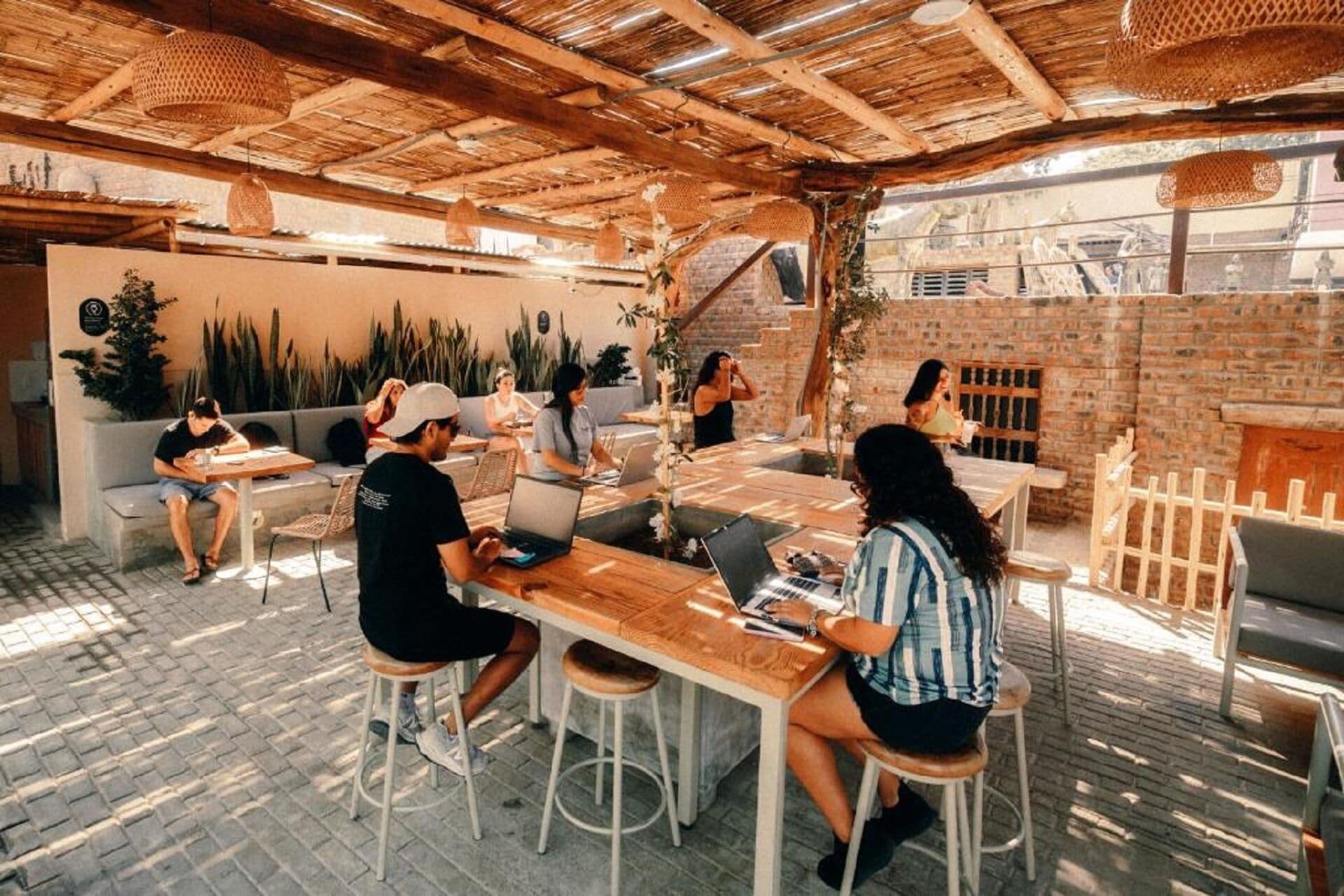 Un café al aire libre con mesas y sillas de madera, rodeado de plantas y un muro de ladrillo.