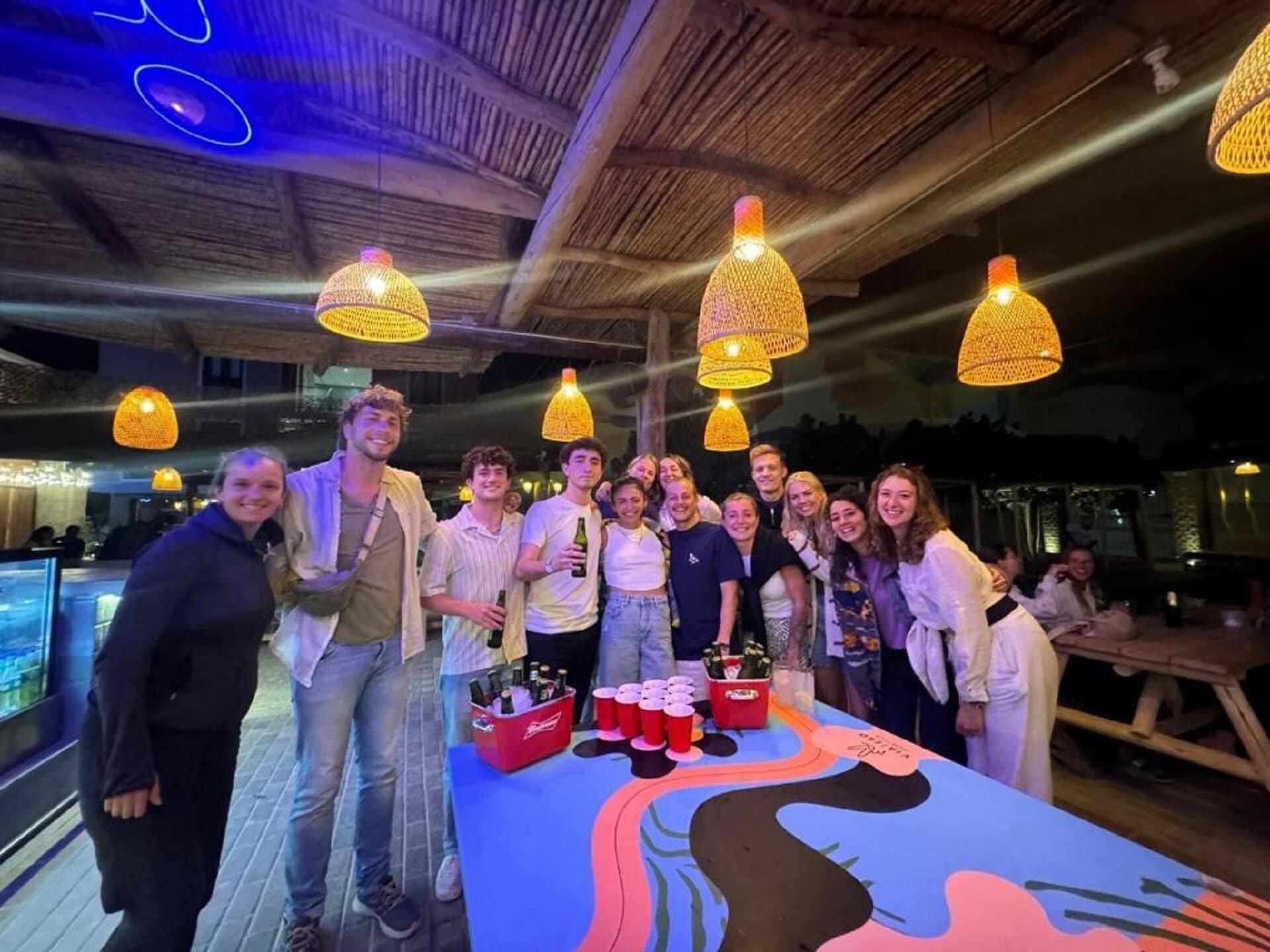 A group of people gathered around a table with a colorful tablecloth, under hanging lamps, in a room with a wooden ceiling.