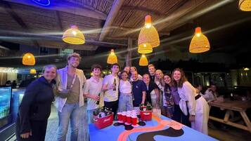 A group of people gathered around a table with a colorful tablecloth, under hanging lamps, in a room with a wooden ceiling.