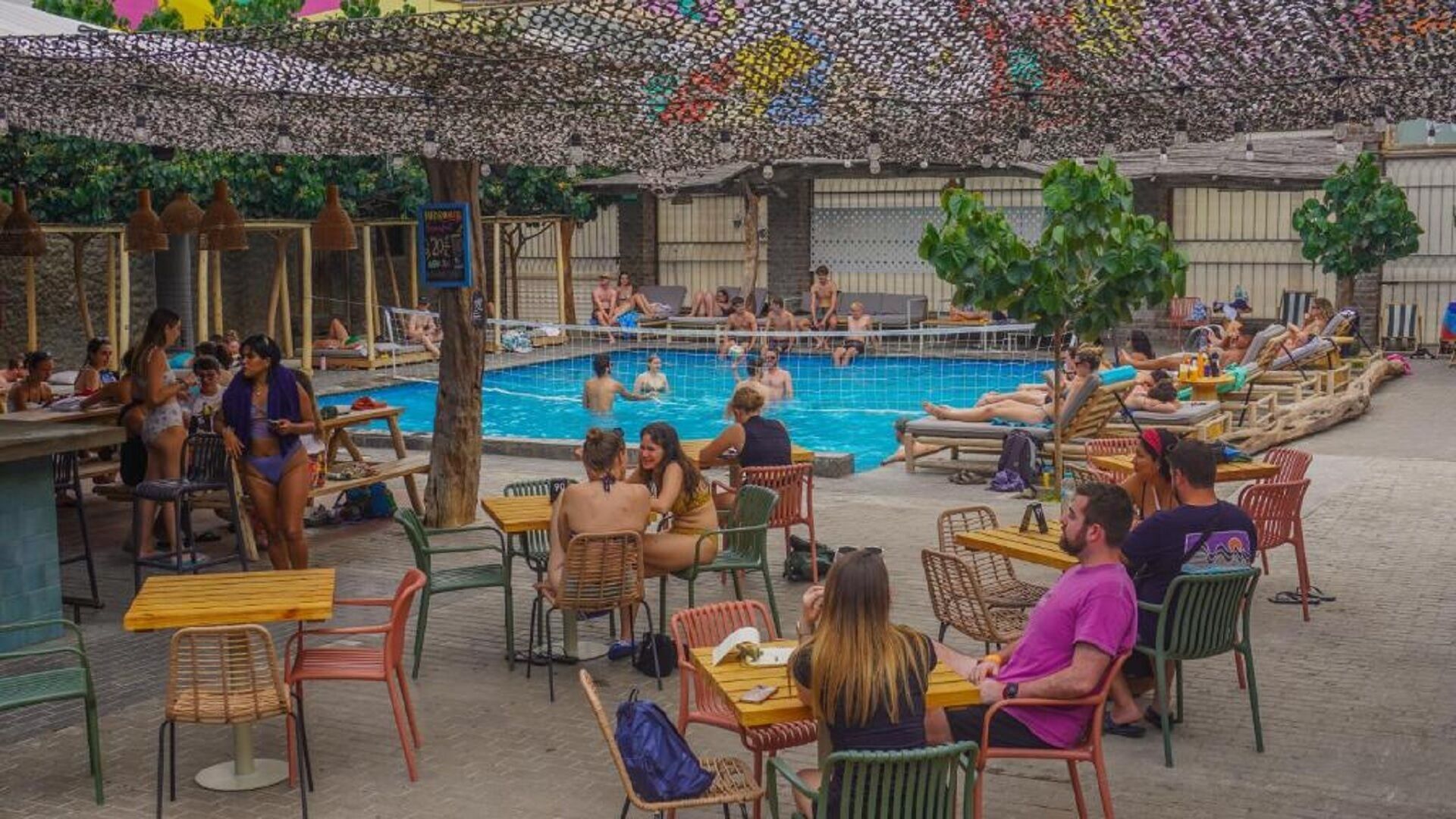 Zona de piscina al aire libre con gente nadando y relajándose, rodeada de mesas y sillas.