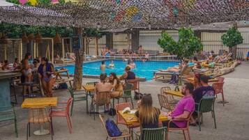 An outdoor pool area with people swimming and relaxing, surrounded by tables and chairs.