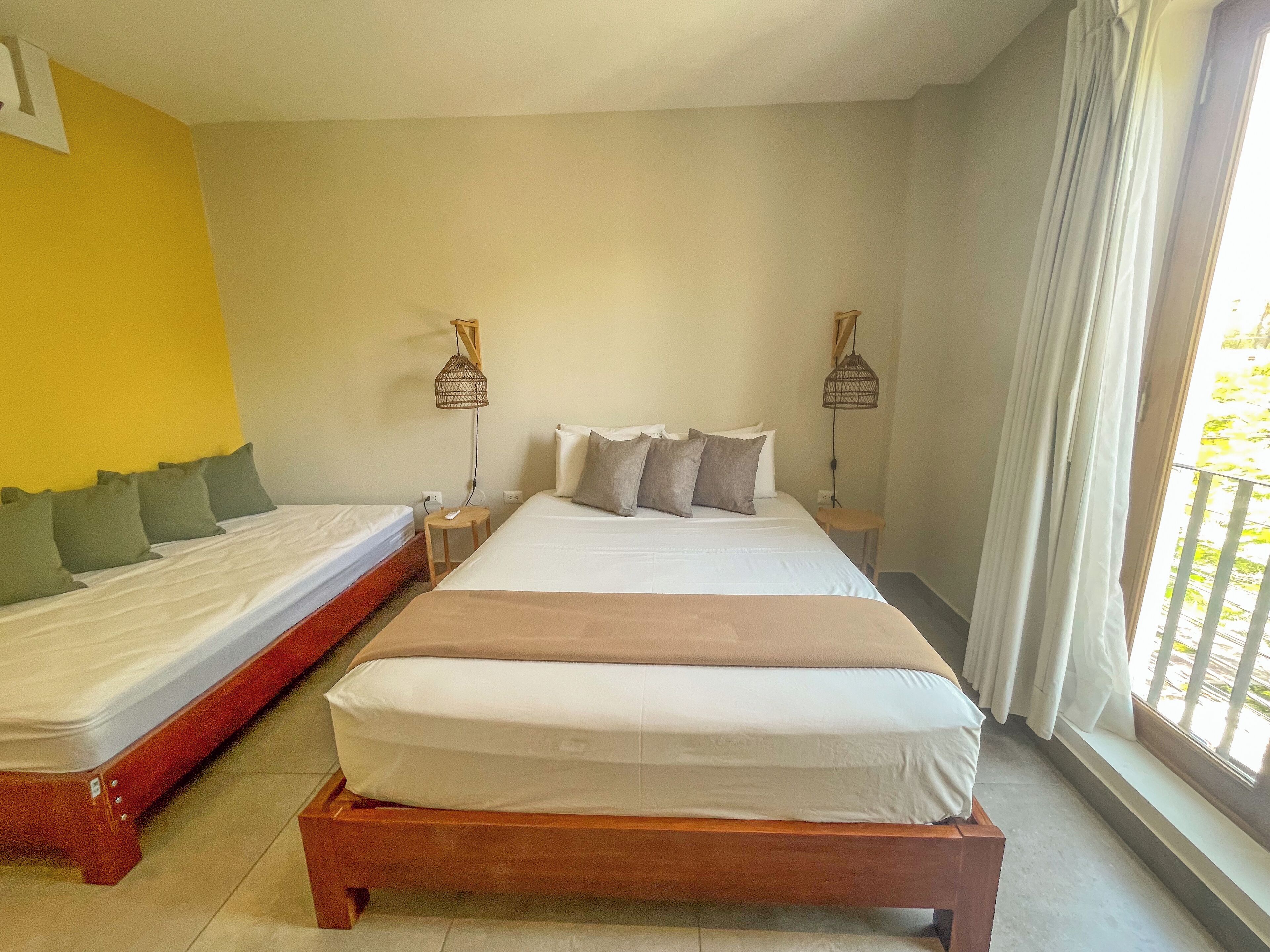 A hotel room with two beds, a wooden headboard, a bedside table with a lamp, and a window with curtains.