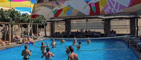 Una piscina con gente en el agua y en el borde, rodeada de palmeras y un mural colorido.
