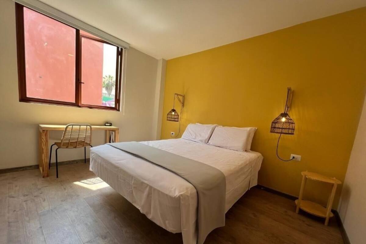 A bedroom with a bed, a desk, and a chair. There is a window with blinds, a wall-mounted lamp, and a small shelf.