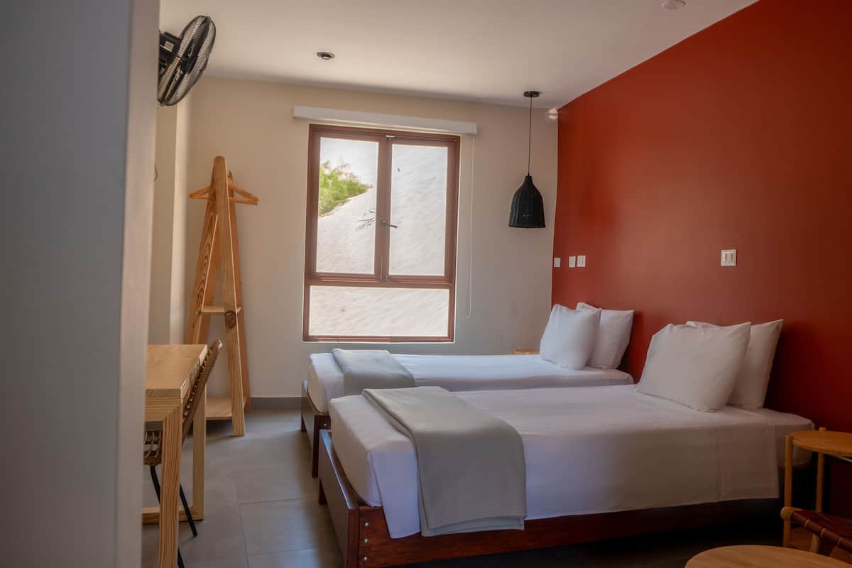 A bedroom with two beds, a wooden desk, a hanging light, and a window with a view of greenery.