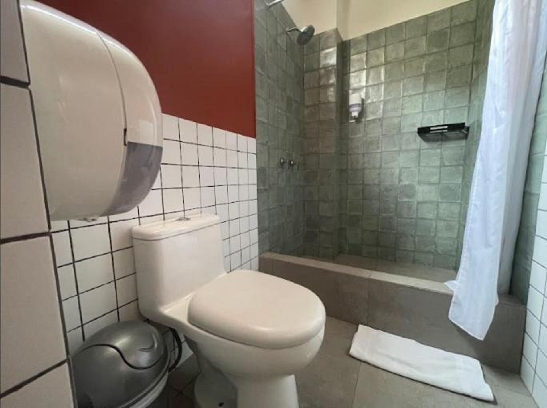 A bathroom with a toilet, a trash bin, a shower with a glass door, and a towel on the floor.