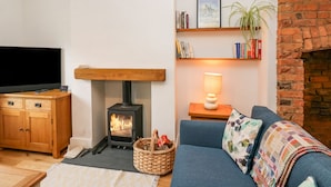 A cozy living room with a wood-burning stove, a television, a bookshelf, and a brick fireplace. - Bay Tree Cottage (Richmond)