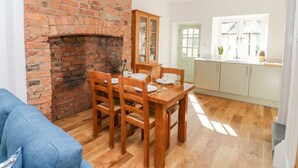 A dining area with a brick fireplace, wooden dining table, and chairs. There is a kitchen area with white cabinets and a window. - Bay Tree Cottage (Richmond)