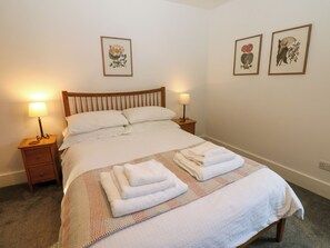 Un lit bien fait, avec du linge de lit blanc et des serviettes pliées, deux tables de chevet avec des lampes, et deux reproductions botaniques encadrées accrochées au mur.
