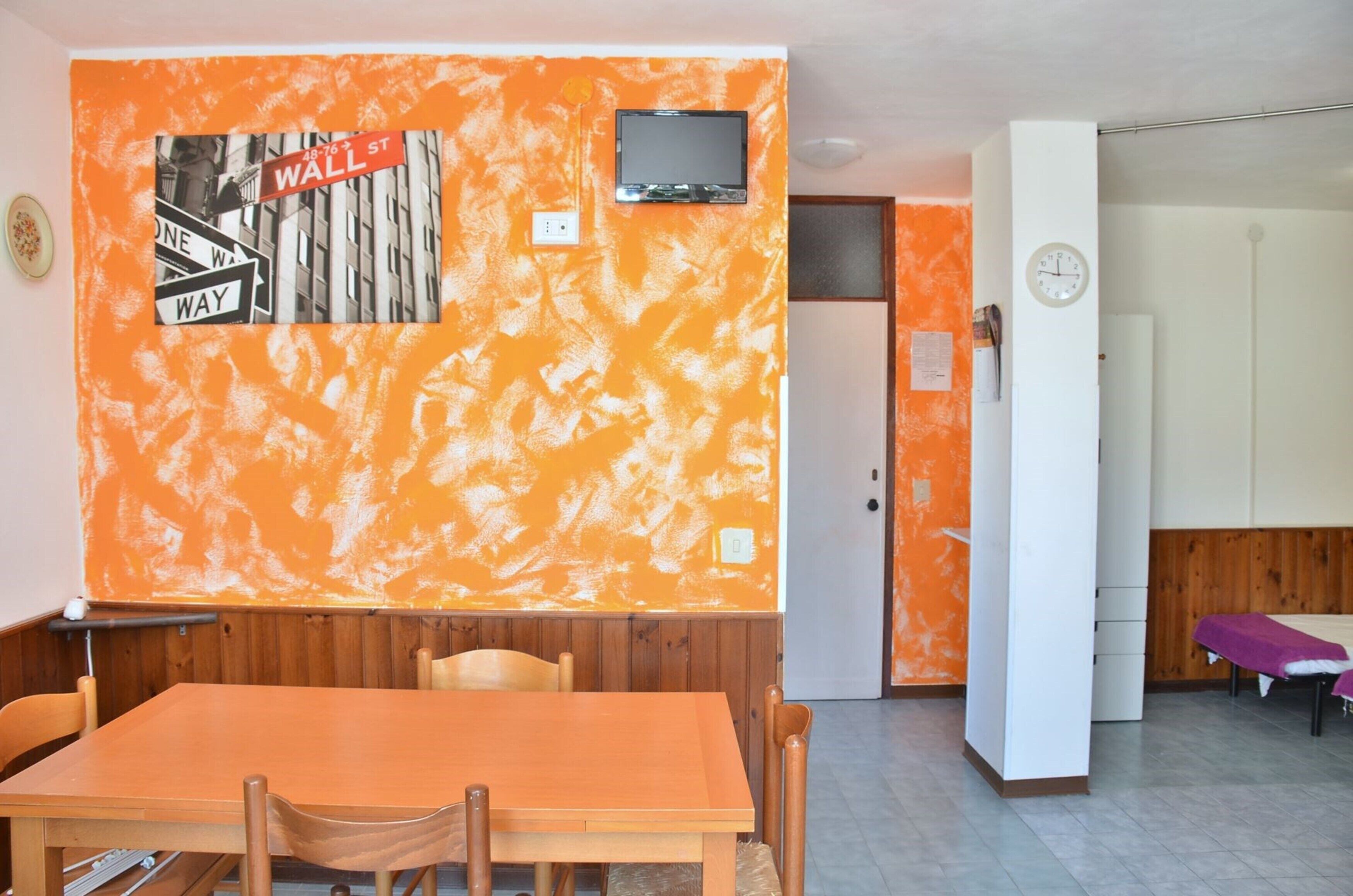Una habitación con una mesa y sillas de madera, una pared naranja con textura y un letrero que dice 'Wall St', un televisor y un reloj en la pared.