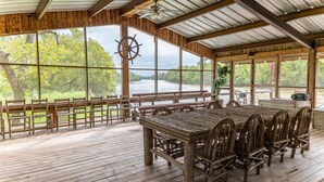 A rustic wooden deck with a large table and chairs, overlooking a river.