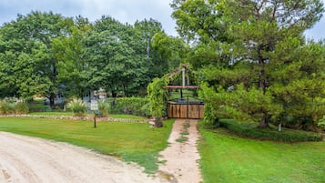 Eine rustikale Holzbrücke über einen Schotterweg, der zu einem Holztor führt, umgeben von üppigem Grün und Bäumen.