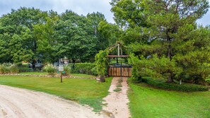 A rustic wooden bridge over a gravel path leading to a wooden gate, surrounded by lush greenery and trees.