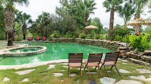 Ein Pool mit einer Steinmauer und einem Reetdachschirm, umgeben von Palmen und Liegestühlen.