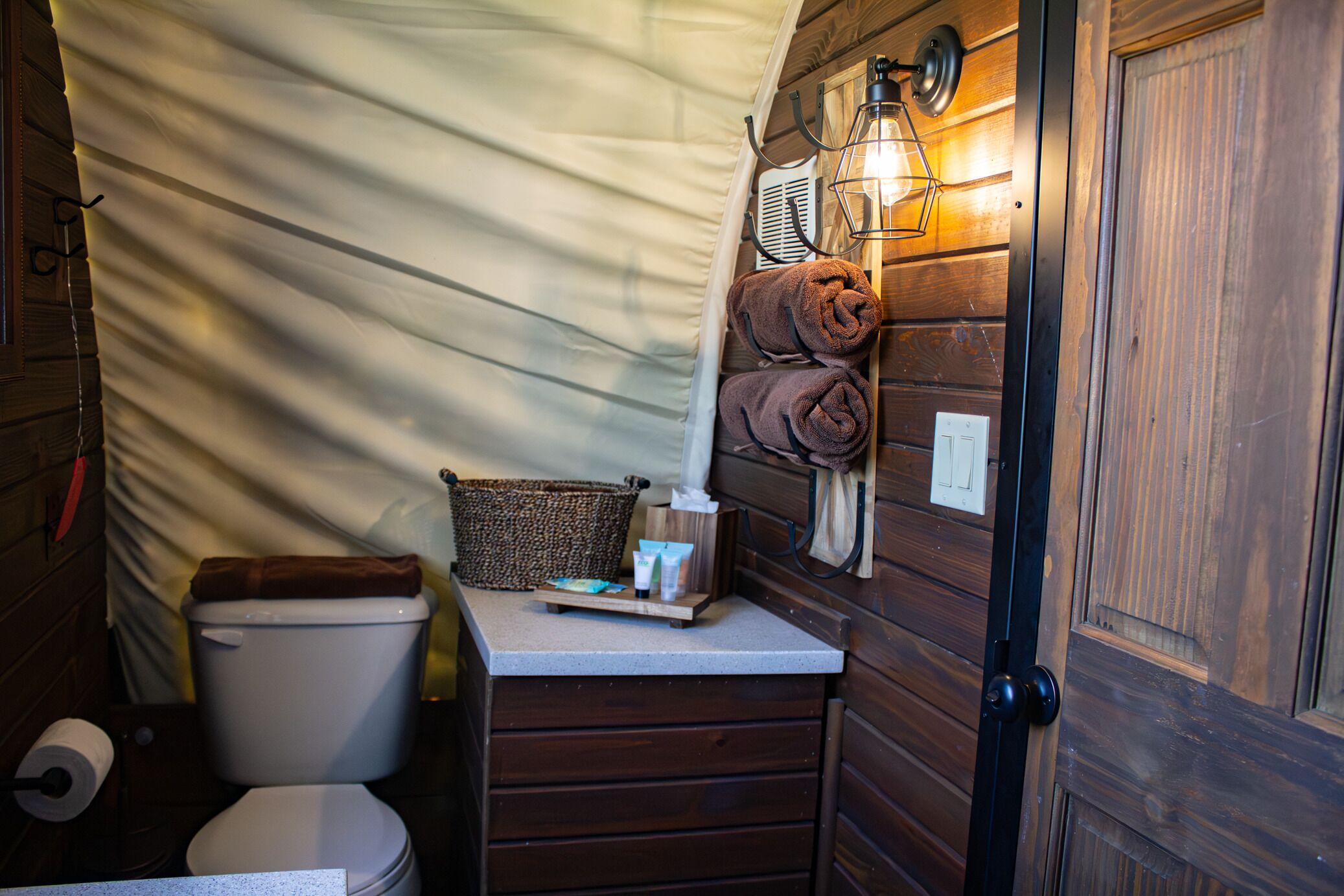 A compact bathroom with a toilet, a towel rack, a basket, and a wall-mounted light fixture.